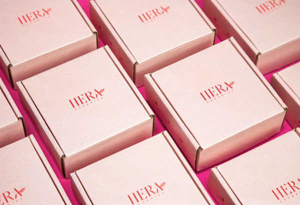 Campaign photography for Hera Organics showing branded product packaging arranged in a geometric pattern on a pink background